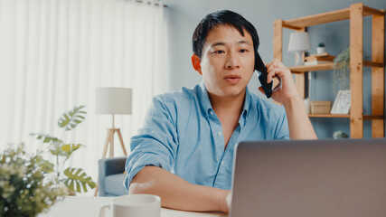 Freelance Asia guy casual wear using laptop talk on cell phone in living room at house. Working from home, remotely work, distance education, social distancing, quarantine for corona virus prevention.