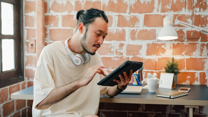 Freelance Asia guy using tablet for searching data and social media on internet in living room at...