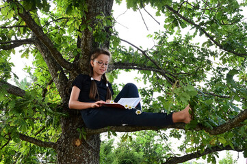 cute teenage girl with glasses is sitting on branch of large tree, an oak in summer park and reading a book. Concept of training and education. Back to school