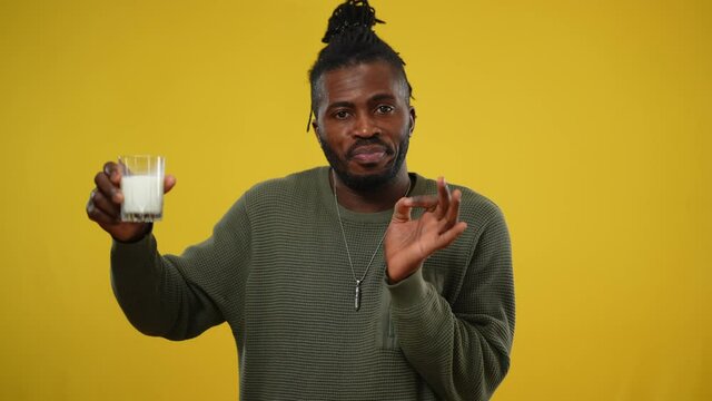 Dissatisfied African American man smelling and tasting milk making disgust facial expression. Portrait of handsome guy posing with dairy in glass at yellow background. Lactose intolerance