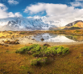 Manaslu mountain in clouds and lake at sunrise in Nepal. Beautiful landscape with snowy mountains, green bush, mountain lake, hill, reflection, grass, blue cloudy sky, people. Rocks in snow. Himalayas