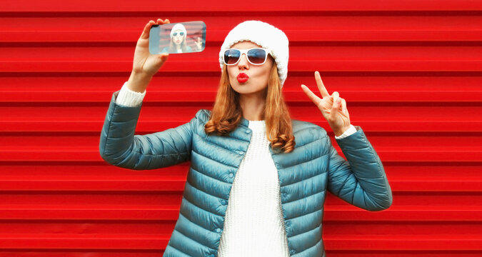 Portrait of stylish young woman taking selfie by phone blowing her lips wearing a white hat, jacket on red background