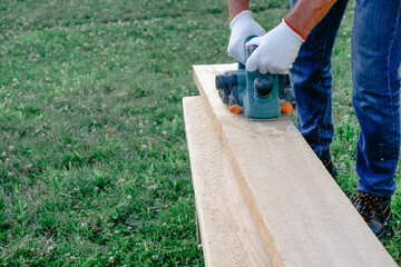 A male joiner planes a wooden plank on the lawn with an electric plane.