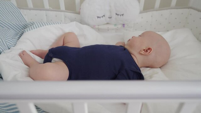 Cute 3 months old baby boy sleeping in a crib under dream cloud lullaby night light, infant in white baby cot closeup. High quality 4k footage