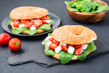 Delicious ready-to-eat bagels stuffed with tomatoes, feta and spinach leaves on a slate board on a black background. Light healthy snack