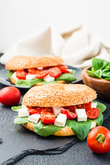 Delicious ready-to-eat bagels stuffed with tomatoes, feta and spinach leaves on a slate board on a black background. Light healthy snack. Vertical view