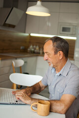 Trendy mature man is working from home with laptop sitting at the table in his kitchen, happy retirement, home-office concepts