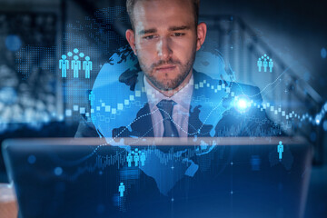 Handsome businessman in suit at workplace working with laptop to hire new employees for international business consulting. HR, social media hologram icons over office background