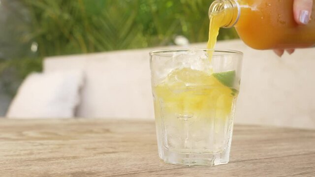 a girl with a beautiful manicure on a sunny day pours orange jamu a natural antibiotic into a glass glass with ice. A close-up of a glass standing on a table in which juice is poured. Slow motion.