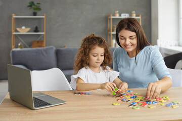 Fototapeta premium Smiling mother or family tutor helping small child learn foreign language. Happy woman and little girl sitting at desk with laptop and developing English reading skills through fun engaging activities