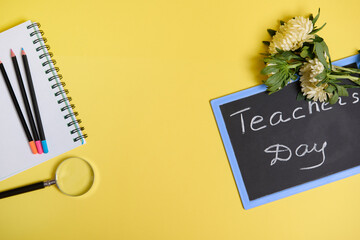 Flat lay of asters flowers lying down on chalkboard with text Teachers Day , and a magnifier loupe next to colored pencils on a blank sheets of a notepad isolated on yellow background with copy space