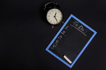 A chalk board with lettering list to do, with empty copy space and a vintage alarm clock. Isolated on black background. Planning concepts, time management, timetable and timing.