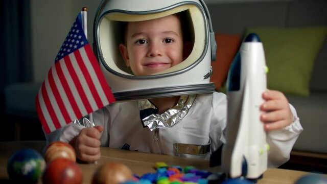 Child playing at home in an astronaut, portrait of a little boy 5 years old in an astronaut costume, smiling happy child looking at the camera,close-up, pilot holding an American flag,travel to space