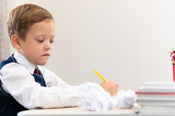 A cute boy first grader in a school uniform does homework while sitting at a desk with a pencil in his hands at home. Remote education. Selective focus. Close-up. Portrait