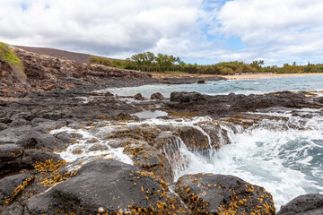 Waves on a rocky reef on a beach with a sandy beach in the distance, in Hawaii