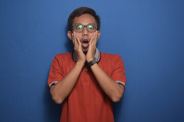 Asian man wearing orange casual tshirt and glasses over blue background afraid and shocked, surprise and amazed expression with hands on face