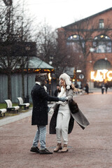 Romantic vacation in X-mas holidays: man and woman in love have fun outdoors enjoy snowfall in old town street. Happy couple in warm fur hats, mittens and coats spin and laugh under snow holding hands