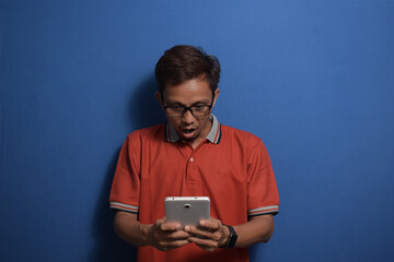 Young Asian man wearing orange casual t-shirt and glasses shocked with a surprise face at the good news he received from histablet