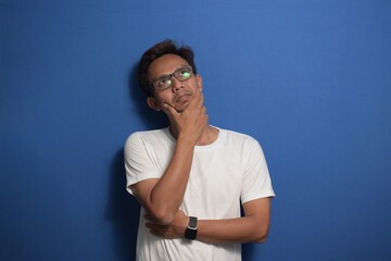 Fototapeta premium Asian man wearing white t-shirt thinking and looking up over on blue background with hand on chin.