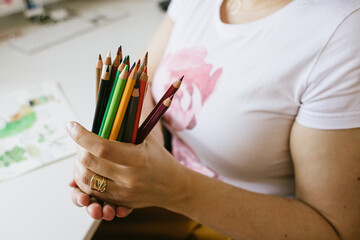 a bunch of colour pencils in woman's hands , a desk with drawings in background - 452593549