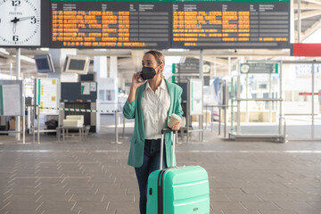 Caucasian girl waits for her train at the station while talking on the phone. Business woman with mask. Concept travel, business and pandemic