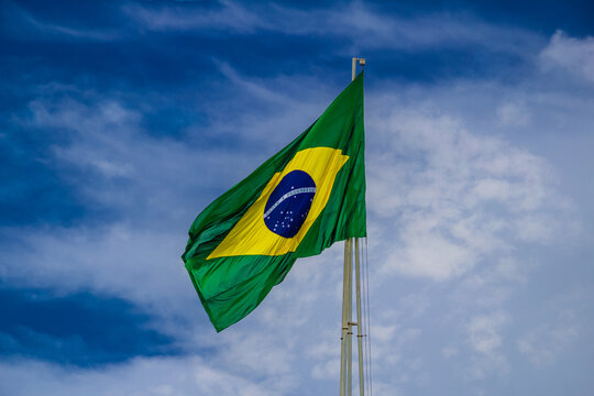 Bandeira do Brasil hasteada, tremulando ao vendo e com céu azul e algumas nuvens ao fundo.