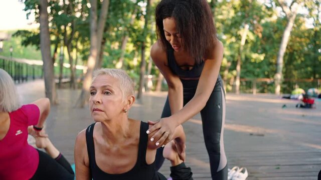 Professional black woman coach helping senior lady with stretching exercise during group sport practice outdoors