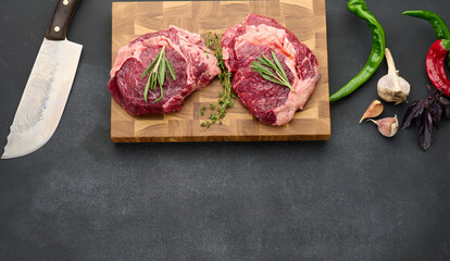 raw piece of beef ribeye with rosemary, thyme on a black table, top view