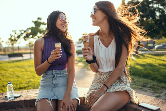 Two beautiful young women eating ice cream while having fun walking through the park in the city.