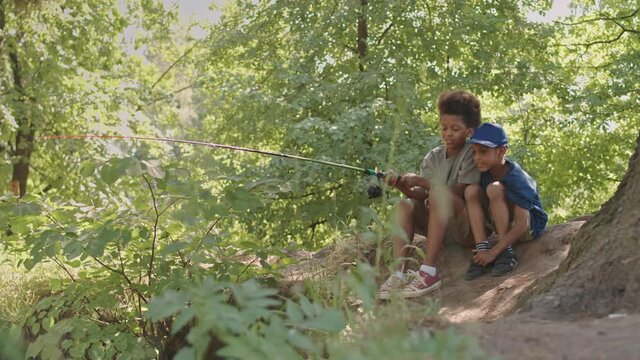 Slowmo shot of African American siblings in casualwear with backpacks fishing together sitting by pond or river in forest on warm sunny day