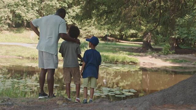 Rear-view slowmo shot of African American man and his two little sons standing by pond in beautiful summer forest fishing