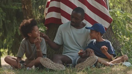 Slowmo shot of cheerful African American man and his cute little sons resting under big tree with hanging American flag on it, eating apples and chatting during summer camping trip - Powered by Adobe