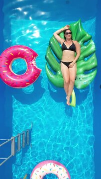 TOP DOWN, AERIAL, VERTICAL: Caucasian female in a black bikini relaxes in her garden pool on a sunny summer weekend. Beautiful young woman lies on a big leaf floatie in her crystal clear home pool.