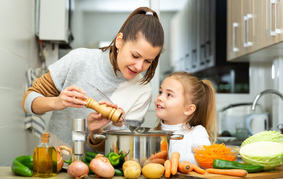 Little daughter helping cooking soup and mother add pepper to pan