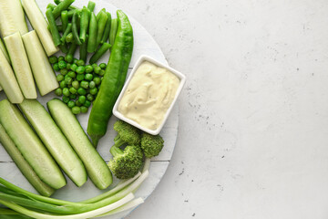 Board with different green vegetables and bowl with sauce on light background