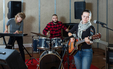 Fototapeta na wymiar Portrait of active excited cheeful brunette girl rock singer with guitar during rehearsal with male drummer and female keyboardist in studio