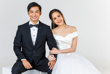 Young attractive Asian couple, soon to be bride and groom, woman wearing white wedding gown. Man wearing black tuxedo, sitting down together. Concept for pre wedding photography