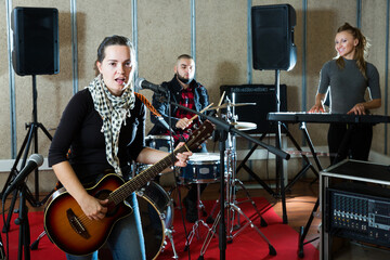 Fototapeta na wymiar excited adult girl rock singer with guitar during rehearsal with male drummer and female keyboardist in studio