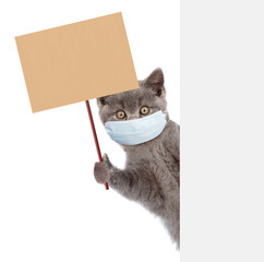 Kitten wearing medical protective mask look from behind empty banner and holds placard on wooden stick. Isolated on white background