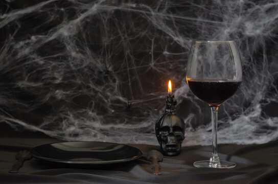 Scary Halloween Dinner Party Concept With A Glass Of Red Wine, Black Plate, Knife, Fork And Skull Candle With Cobweb On Black Background.