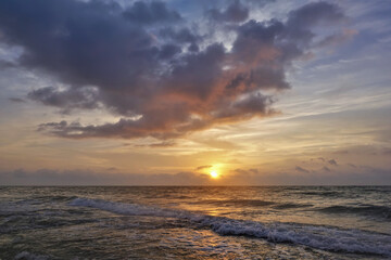 Dawn over the Caribbean Sea. The sun is low on the horizon. There are mauve clouds in the sky. The surf waves are foaming on the sandy beach. A sunny path on the water. Mexico