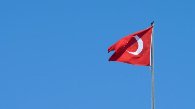 Flag of Republic of Turkey swings in wind. Red field with white star and crescent slightly left of center. Country straddling Western Asia and Southeast Europe. Wind blows on blue sky background