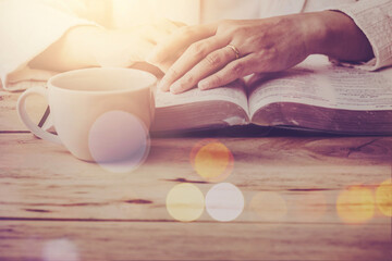 Obraz na płótnie Canvas Close up of a woman hand prays while reading the open bible, blurred page with a coffee cup on wooden table with window light , Christian prayers or bible study concept background with copy space