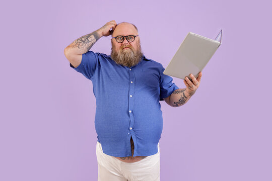 Doubting middle aged man with overweight in tight blue shirt scratches head holding laptop as book on purple background in studio