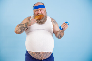 Funny plus size man eats chicken leg and does sports exercises with dumbbell standing on light blue...