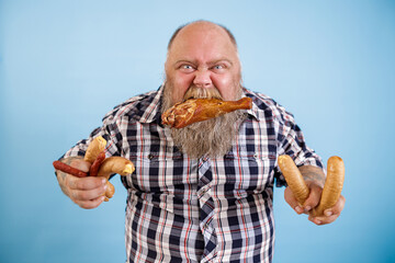Greedy mature obese man holds smoked chicken leg in teeth and bunches of sausages on light blue background in studio