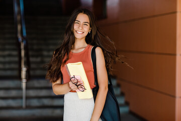 Portrait of a charming student girl with a smile, holding a folder, glasses and a phone
