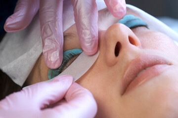 Cosmetologist applying eye patch under client's woman eye on lifting lash procedure in cosmetology clinic. Eyelashes lamination procedure, working process in beuty salon, face of young woman closeup.