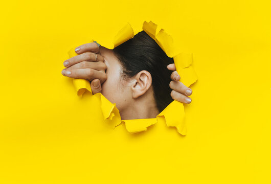 Close-up of a woman's ear and hands through a torn hole in the paper. Yellow background, copy space. The concept of eavesdropping, espionage, gossip and tabloids.