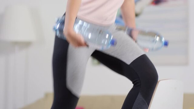 Tilt Up Shot Of Young Woman In Sportswear Doing Step Ups On Chair With Water Bottles As Dumbbells While Training At Home